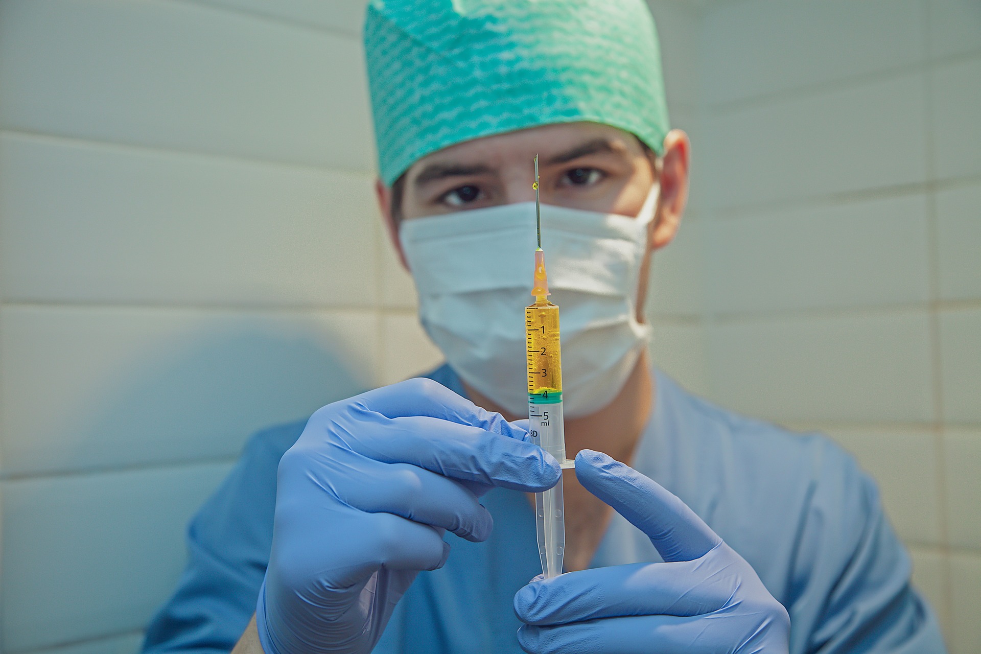 Masked clinician in blue scrubs drawing medication from a vial into a syringe